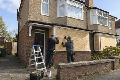 Wide view of a vacant two-storey British semi-detached house with ground-floor windows securely boarded; pair of tradespeople finishing one panel from pavement level, clear afternoon.