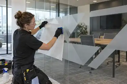 Modern corporate meeting room: woman film installer aligns a custom abstract frosted geometric privacy band on a clear glass partition using transfer film and squeegee, no lettering or logos.